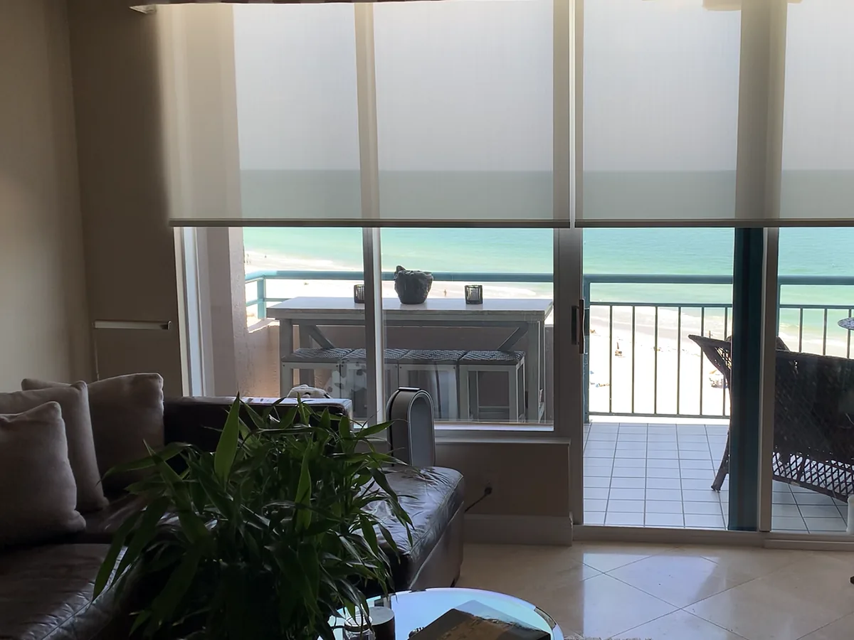 Motorized roll-down solar shades lowered over floor-to-ceiling sliding glass doors in a beachfront Florida living room.